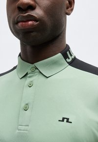 Άνδρας που φοράει ανοιχτοπράσινο πόλο πουκάμισο με τρία κουμπιά και μαύρο λογότυπο στο στήθος, καθώς και μαύρο γιακά με μερικά λευκά γράμματα.