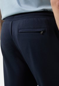 Granatowe spodnie o gładkiej fakturze, z tylną kieszenią ozdobioną małym metalowym detalem oraz elastycznym paskiem w talii dla wygody.
