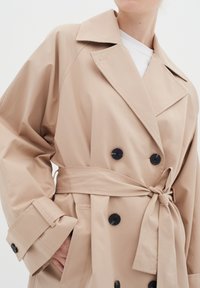 Béžový trench coat vyrobený z hladkej tkaniny, s dvojradovým zapínaním, čiernymi gombíkmi a opaskom na viazanie s širokými manžetami.