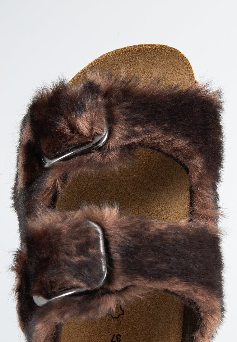 Sandale en fausse fourrure marron avec deux boucles argentées réglables et un lit de pied en tan contourné. Texture douce avec une forme arrondie et un design ouvert.