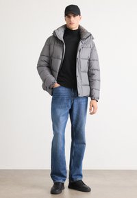 Gray puffer jacket with a high collar and button details, worn over a black turtleneck. Blue straight-leg jeans and black sneakers.