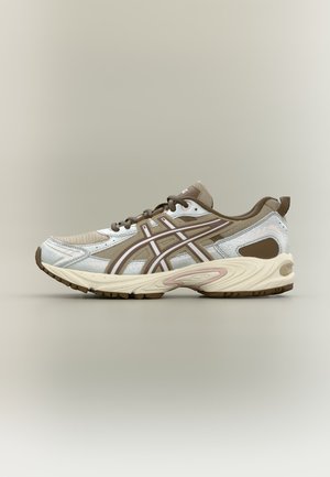 Athletisk sneaker i beige, hvid og brun med mesh-overdel, snørelukning og polstret sål, vist fra ydersiden på en neutral baggrund.