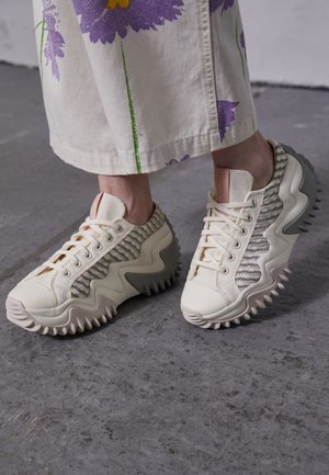 Bílé a šedé tenisky na šněrování s vlnitou podrážkou a texturovanými boky, nošené s krémovou sukní s fialovými a žlutými květinovými vzory.