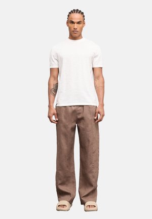 Hombre con el pelo trenzado de pie mirando hacia adelante, vistiendo una camisa blanca de manga corta, pantalones marrones de pierna ancha y sandalias beige deslizantes.