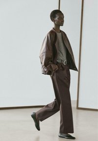 Große Person geht drinnen und trägt eine braune Lederjacke über einem grauen Hemd, weite braune Hosen, schwarze Ballerinas und einen schwarzen Gürtel.