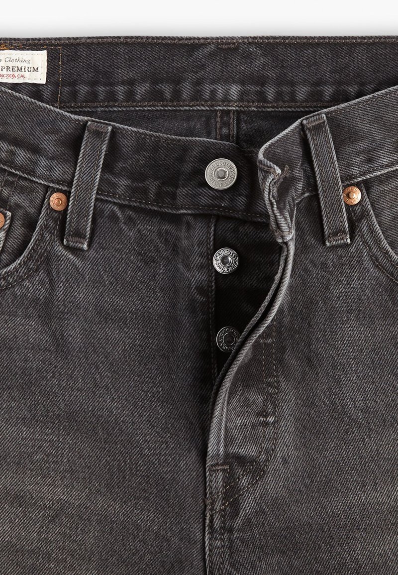 Jean en denim noir à taille haute, avec des accessoires en cuivre et en argent, fermeture à cinq boutons et texture décolorée.