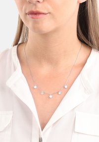 In primo piano, una donna indossa una collana d'argento con cinque pietre preziose rotonde e trasparenti equidistanti su una catena delicata.