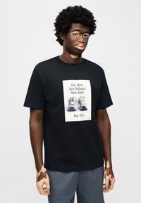 Μαύρο μπλουζάκι με γραφική εκτύπωση αθλητικών παπουτσιών New Balance, με το κείμενο «Το πιο New Balance παπούτσι ποτέ. Το 574.» Γκρι παντελόνι.