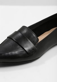 Mocassin en cuir noir à bout pointu avec une large lanière sur le dessus et une semelle intérieure beige légèrement rembourrée.