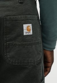 Κοντινό πλάνο στην τσέπη σκούρου γκρι παντελονιού Carhartt με λευκή και πορτοκαλί ετικέτα λογότυπου Carhartt, που φορά ένα άτ ομο με μπλε-πρασινωτό μακρυμάνικο πουκάμισο.