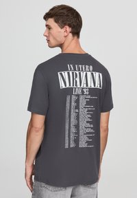 T-shirt grigia in cotone con maniche corte, caratterizzata da un grande testo bianco sul dorso che elenca le date dei concerti del tour "In Utero" dei Nirvana.