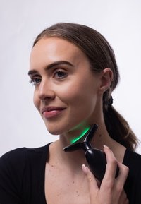 Czarny masażer do twarzy trzymany w pobliżu szyi, z zakrzywionym kształtem i zieloną diodą wskaźnikową. Gładka tekstura z ergonomiczną rączką.