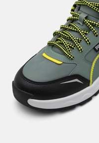 Gray athletic shoe with yellow accent, black rubber sole, textured green upper, and black and green mixed laces. Reflective detailing included.