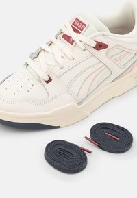 Baskets crème avec bout perforé, semelle contrastante bleu marine et accents de couleur claire. Comprend deux paires de lacets en gris foncé.