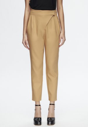 Femme portant un pantalon taille haute camel ajusté avec un détail asymétrique à l'avant et des talons plateformes noirs à bride à la cheville.