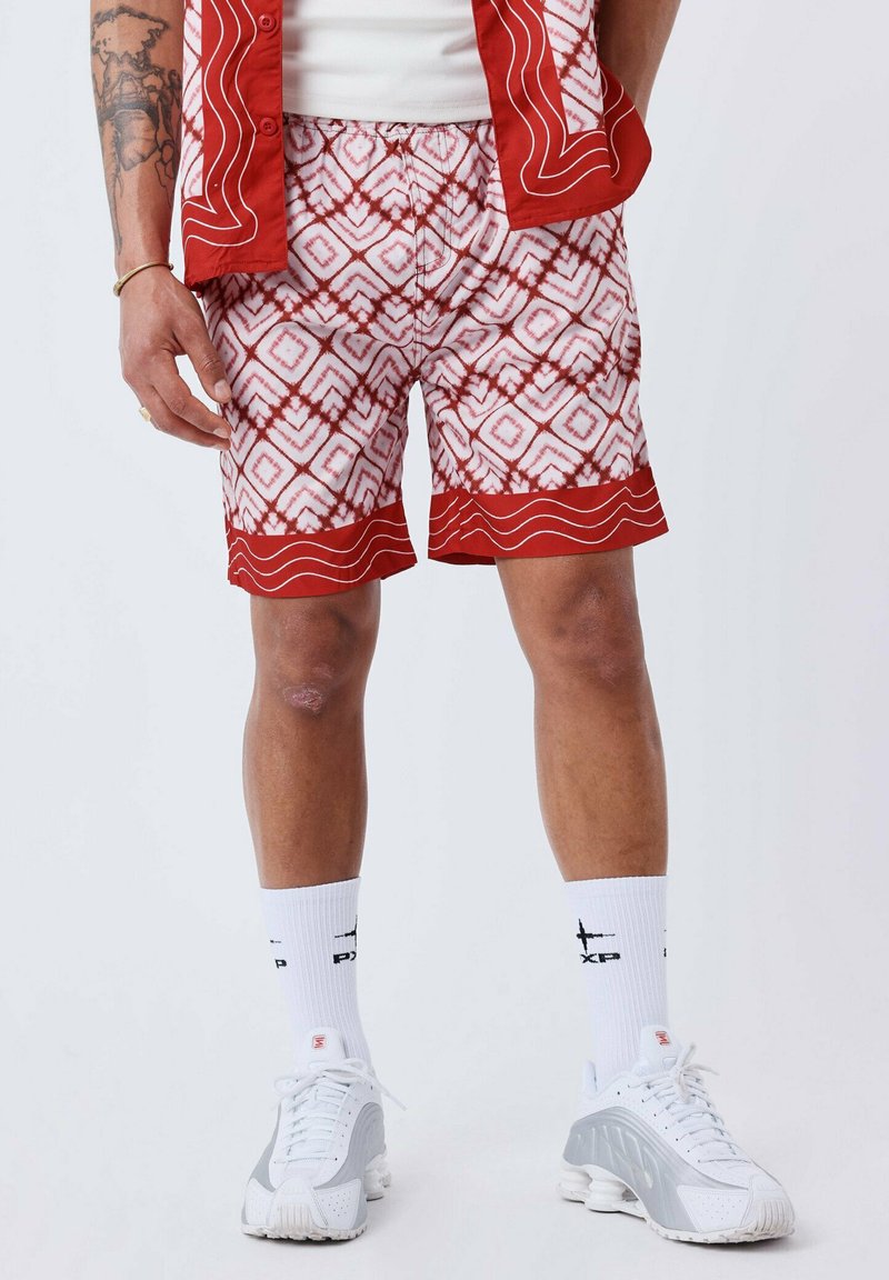 Pantalones cortos de color rojo y blanco con un patrón de formas de diamante, cinturilla roja sólida y detalle de dobladillo ondulado. Combinados con calcetines blancos y zapatillas deportivas grises.