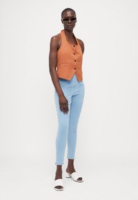 Rustrød, ermeløs vest med frontknapper og to lommer, parret med lyseblå skinny jeans og hvide åbne hæle.