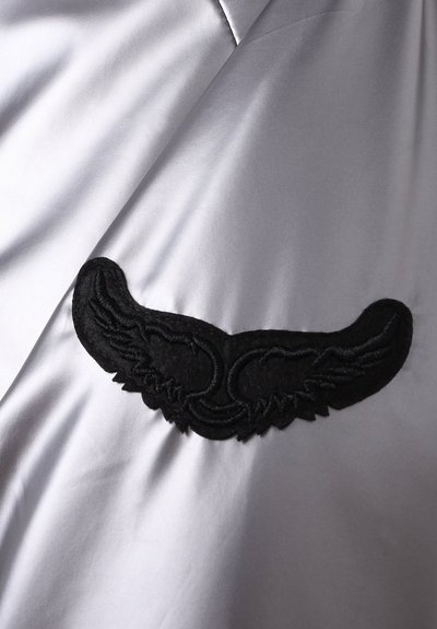 Sølv satengstoff med svart brodert vingemønster. Teksturen er glatt og skinnende, med en fremtredende dekorativ detalj.