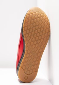 Chaussure en tissu rouge avec une semelle en caoutchouc texturée de couleur beige présentant des motifs hexagonaux, des bordures noires et un logo embossé au bas.