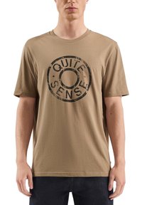 T-shirt en coton marron à manches courtes, avec un imprimé circulaire noir arborant le texte "QUITE SENSE" en police usée, centré sur le devant.