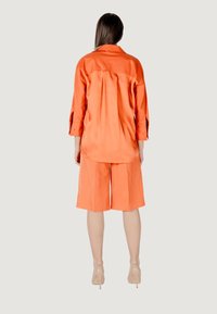 Camicia arancione a maniche lunghe con bottoni, abbinata a pantaloni corti arancioni fino al ginocchio. Tessuto morbido, vestibilità rilassata e dettagli discreti sulla cucitura posteriore.