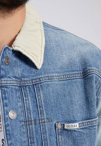 Denim jacket with light blue wash, corduroy collar, silver hardware, and a small "GUESS" label on the left pocket.