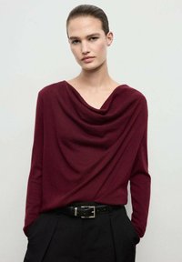 Burgundy draped top in soft knit material, featuring a cowl neckline, long sleeves, and a relaxed fit, paired with black tailored trousers.