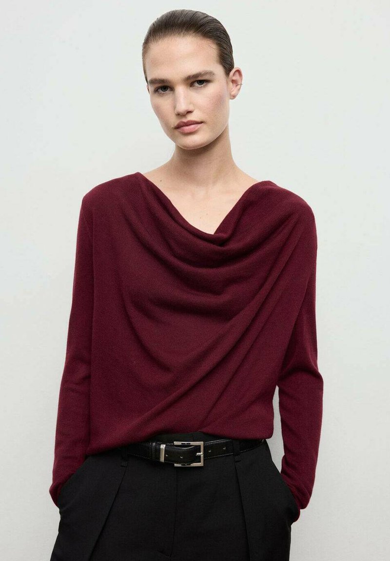 Burgundy draped top in soft knit material, featuring a cowl neckline, long sleeves, and a relaxed fit, paired with black tailored trousers.