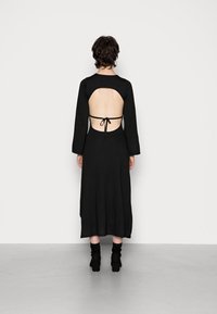 Abito nero a maniche lunghe con schiena scoperta e dettaglio annodato, realizzato in tessuto morbido con una silhouette fluida e orlo fino alla caviglia.