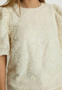 Cream-colored top with a textured floral pattern, featuring short sleeves and a round neckline. Fabric appears soft and slightly plush.