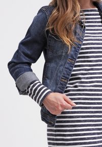 Veste en denim au coupe classique, avec un délavage bleu foncé, des poignets gris contrastants et des accents usés ; portée sur une robe rayée gris et blanc.