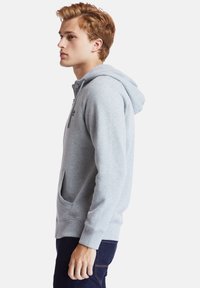 Felpa grigia chiara con cappuccio, chiusura con zip, tasca a marsupio frontale, polsini a costine e un piccolo logo sul petto.