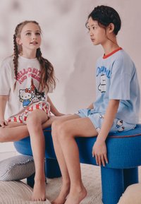 Deux enfants sont assis sur un tabouret rembourré bleu. L'un porte un t-shirt Hello Kitty blanc avec un texte rose et un short à carreaux rouges ; l'autre porte un t-shirt bleu avec des accents rouges et un short bleu clair avec de petits motifs Hello Kitty.