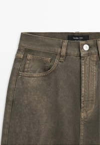 Jean en denim vert olive à taille haute, avec un détail de poche avant et une étiquette logo. Présente des coutures visibles et un bouton métallique à la braguette.