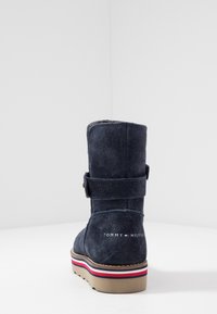 Ciemnoniebieski zamszowy botek za kostkę z detalem w postaci paska, logo na tyle oraz wielokolorową paskowaną podeszwą w kolorze czerwonym, białym i niebieskim.