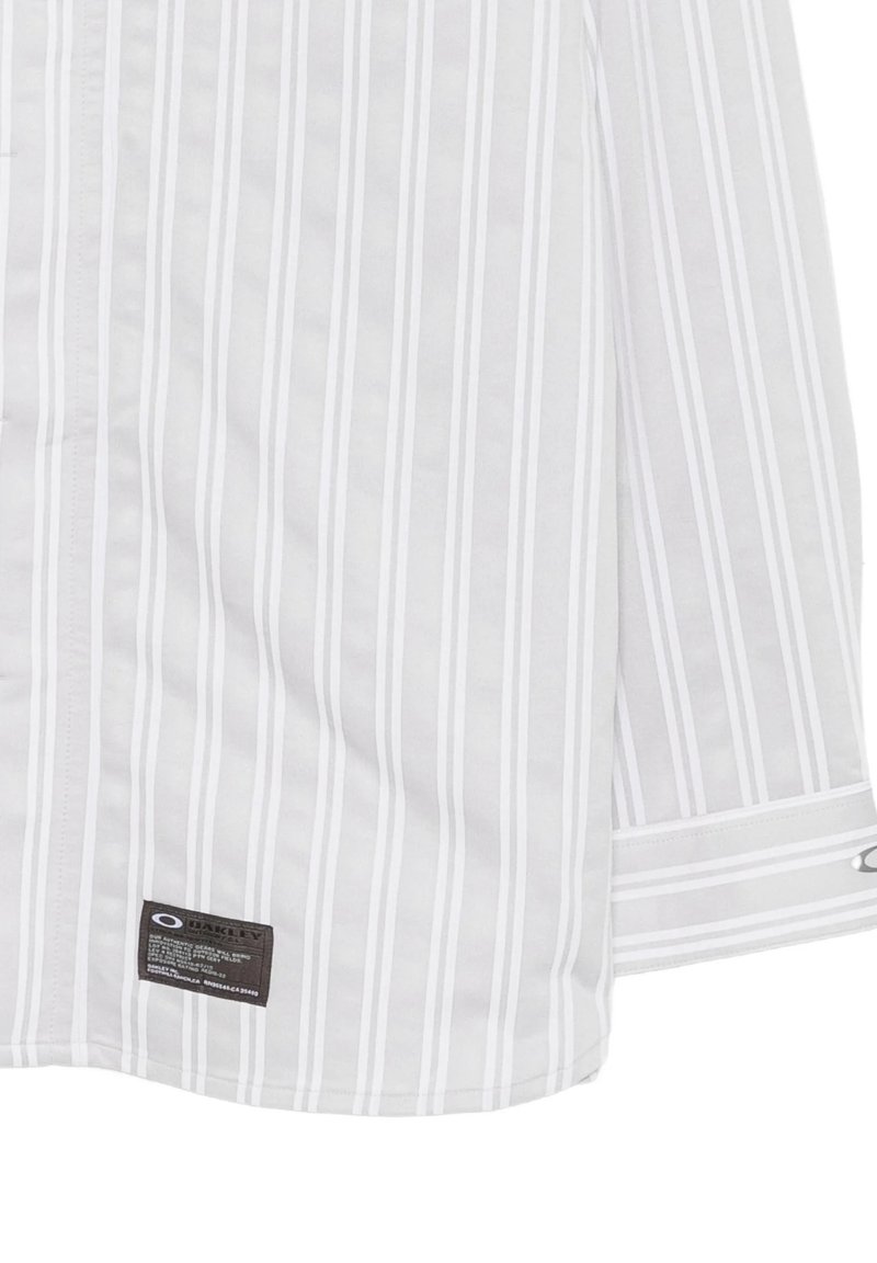 Camicia bianca con strisce verticali grigio chiaro e una piccola etichetta nera del marchio Oakley vicino all'orlo, che mostra parte di un polsino e di una manica.