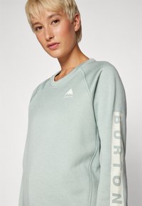 Sudadera verde claro con cuello redondo acanalado, que presenta un logo de montaña blanca y un gráfico de "BURTON" en la manga. Textura suave.