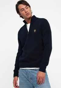 Pulover bleumarin cu guler înalt, închidere cu fermoar, textură corduroy și o mică etichetă cu logo pe piept. Purtați peste o cămașă albă.