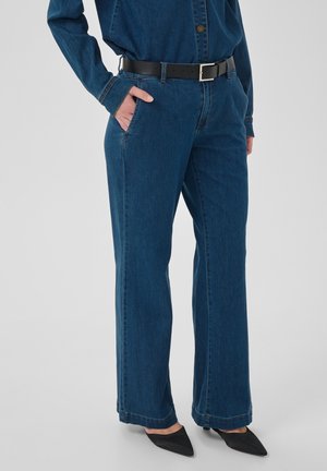 Personne portant un jean en denim bleu foncé à jambes larges avec une ceinture noire et des chaussures noires à bouts pointus, main dans la poche, sur fond uni.