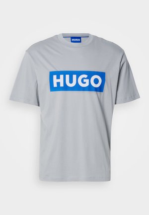 Lys grå kortermet t-skjorte med et stort blått rektangulært panel med hvit tekst "HUGO" over forsiden. Laget av bomull, med rund hals.