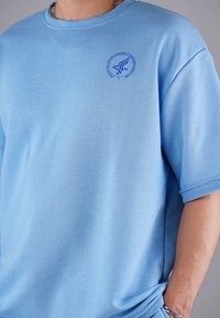 Light blue, short-sleeved t-shirt made of soft fabric, featuring a small blue graphic logo on the left chest and a relaxed fit.