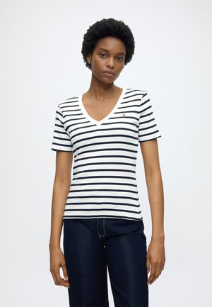 Žena s krátkymi kučeravými vlasmi, ktorá má na sebe biele tričko s výstrihom do V s čiernymi horizontálnymi pruhmi a tmavé džínsy, stojaca pred jednoduchým bielym pozadím.