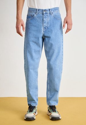 Homme portant un jean droit bleu clair et des baskets multicolores, debout sur un sol jaune devant un fond uni.