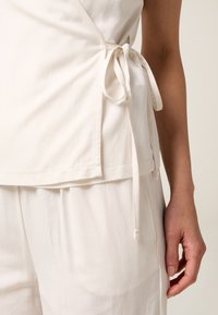 Top avvolgente bianco realizzato in tessuto leggero, caratterizzato da un dettaglio di legatura laterale, texture liscia e vestibilità comoda, abbinato a pantaloni coordinati.