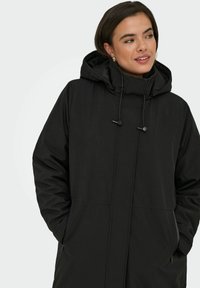 Svart lång jacka med huva, med rak skärning, dragsko-detaljer och sidofickor. Slätt tyg med matt finish.