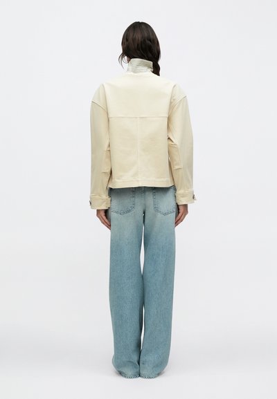 Άτομο με κρεμ μπουφάν με ψηλό γιακά και ανοιχτό μπλε τζιν παντελόνι φαρδιάς γραμμής, που στέκεται γυρισμένο προς τα πίσω μπροστά σε απλό φόντο.