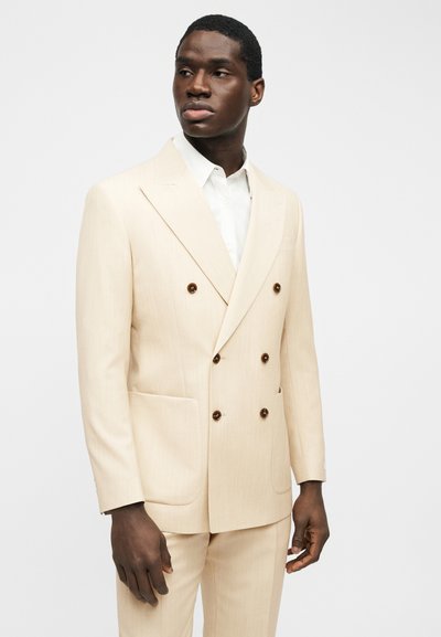 Άνδρας που φοράει μπεζ διπλόκαμπτο κοστούμι με καφέ κουμπιά και λευκό πουκάμισο, φωτογραφημένος κοιτώντας ελαφρώς προς το πλάι σε απλό φόντο.