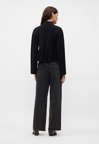 Svart kort genser med peplum-kant, kombinert med høy midje og vide, mørkegrå jeans. Modellen har på seg enkle svarte hæler.