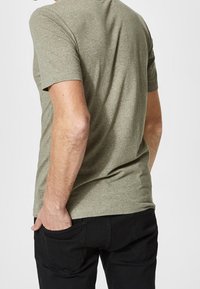 Olivgrön t-shirt med korta ärmar och avslappnad passform, visad bakifrån, i kombination med svarta byxor. Mjuk textur, minimalistisk design.