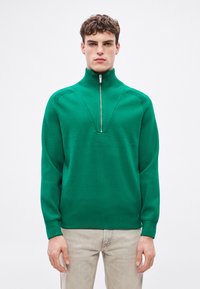 Zielony sweter typu pulower z wysokim kołnierzem i półzamkiem, z prążkowanymi dodatkami i luźnym krojem. Miękka, dzianinowa faktura.